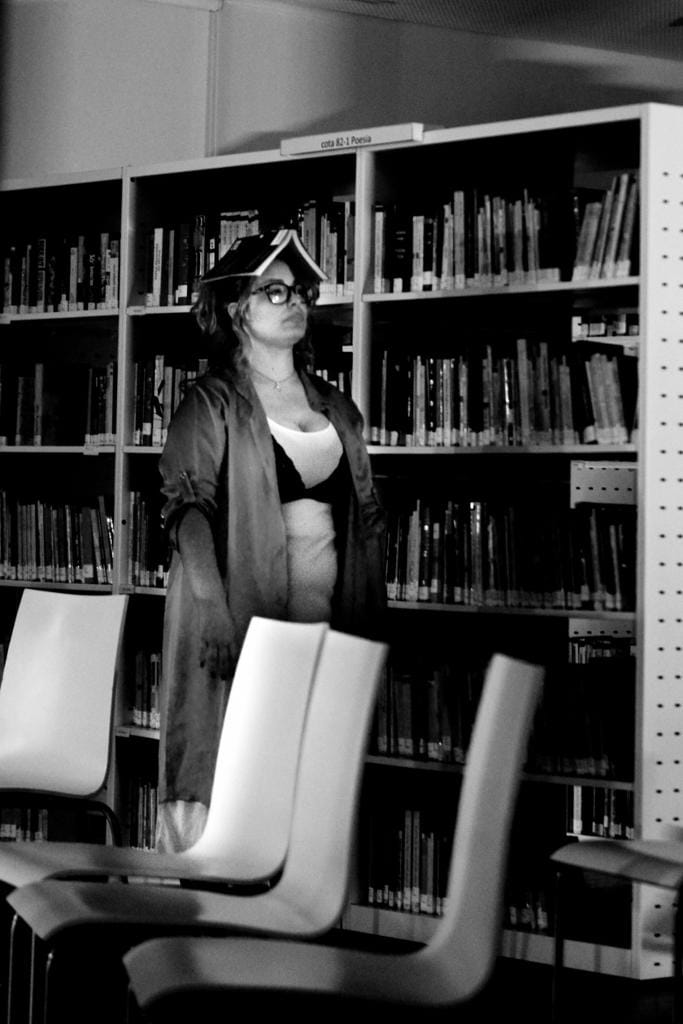 Participante com livro de poesia sobre a cabeça, entre estantes da biblioteca